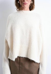 Femme portant un pull en grosse maille crème surdimensionné avec un ourlet et des poignets côtelés, associé à un pantalon taille haute à carreaux marron.