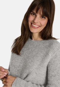 Grauer Strickpullover mit strukturiertem Muster, gerippten Bündchen und Rundhalsausschnitt. Das Modell hält einen Ring und zeigt die lässige Passform und das weiche Material.