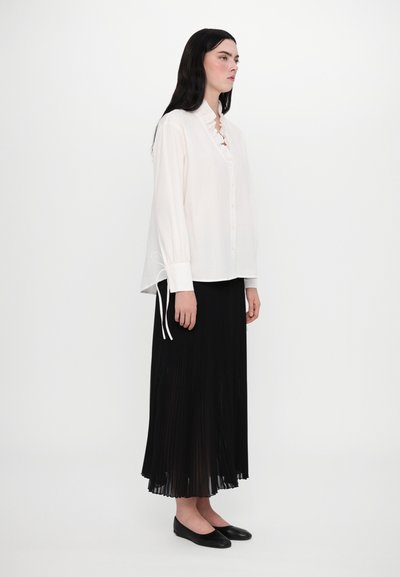 Weiße, knöpfbare Bluse mit Rüschenausschnitt und Bindebändern an den Ärmeln, kombiniert mit einem langen, plissierten schwarzen Rock und schwarzen Ballerinas.