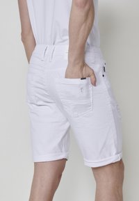 Pantalones cortos de mezclilla blanca con dobladillos enrollados, que presentan costuras visibles, dos Bolsillos traseros y ligeros detalles de desgaste para dar textura.