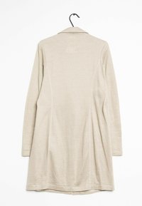 Beżowa kurtka z długim rękawem i kołnierzem, o luźnym kroju, gładkiej fakturze i subtelnych detalach szwów, zawieszona na czarnym haczyku.