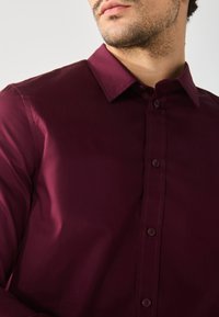 Burgunderfarbene Button-up-Hemd aus glattem Stoff, mit spitzem Kragen, Frontleiste mit passenden Knöpfen und subtiler Schimmer.