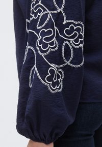 Μπλούζα σε ναυτικό μπλε με λευκή κεντητή διακόσμηση με λουλούδια κατά μήκος του μανικιού, με υφασμάτινη υφή και φουσκωτό μανίκι με ελαστικό τελείωμα.