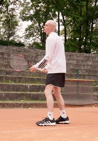Tenisaš u bijeloj majici s dugim rukavima i crnim prugama te crnim šortsem. Drži reket na zemljanoj podlozi s zelenilom u pozadini.