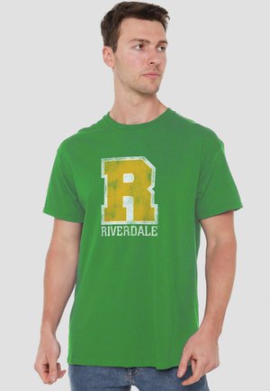 Man wearing green T-shirt with large yellow letter R and word Riverdale, looking to his right against plain background.