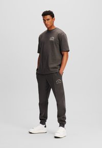 Tricou din bumbac gri, cu mâneci scurte și un logo mic, purtat cu pantaloni de trening asortați, care au buzunare laterale și manșete ribbate. Tenisi albi.