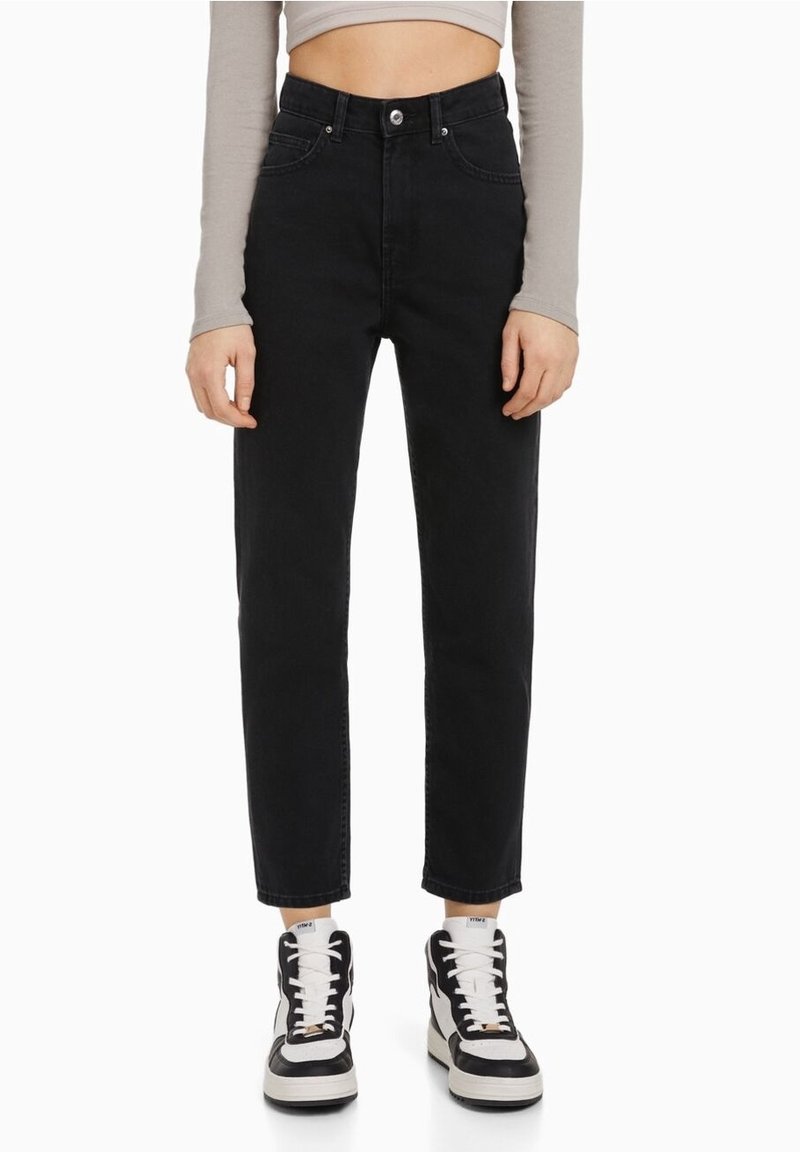Femme portant un jean noir taille haute, des baskets montantes blanches et noires, et un haut gris à manches longues raccourci, debout devant un fond blanc.