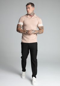 Beige póló, fehér csíkokkal az ujjakon, fekete cargo nadrággal és fehér sportcipőkkel párosítva. A modellen látható tetoválások az karokon.