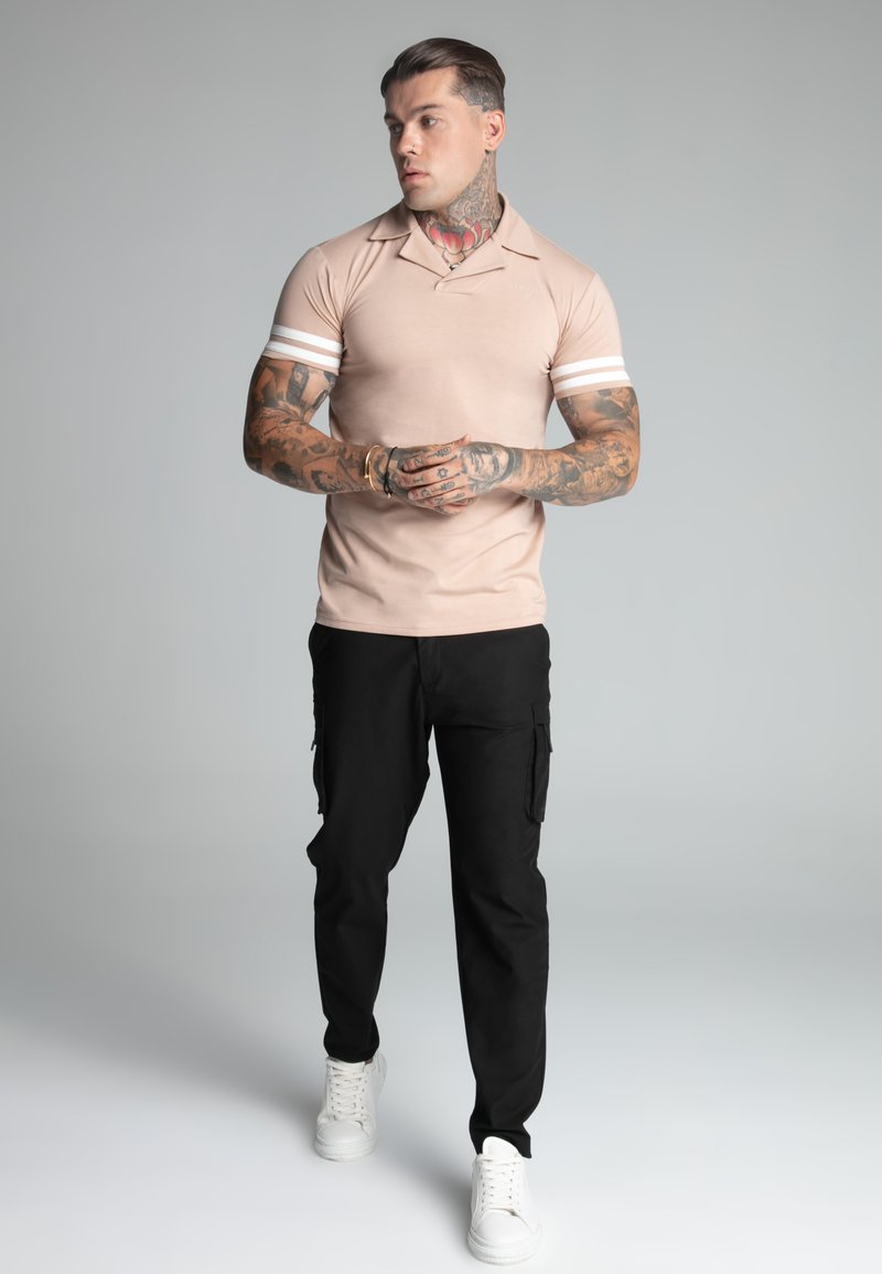 Polo beige con rayas blancas en las mangas, combinado con pantalones cargo negros y zapatillas blancas. El modelo muestra tatuajes visibles en los brazos.