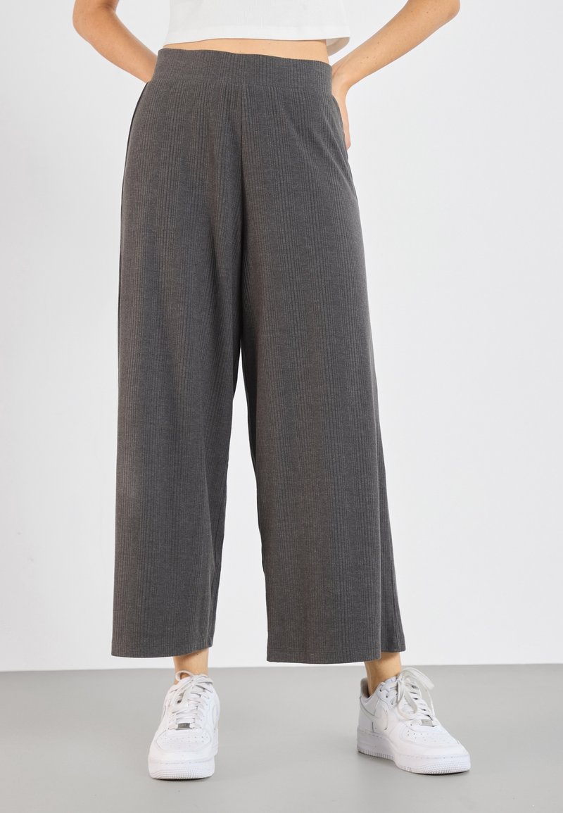 Femme portant un pantalon gris taille haute à jambes larges, associé à des baskets blanches et un top blanc court, debout les mains sur les hanches.