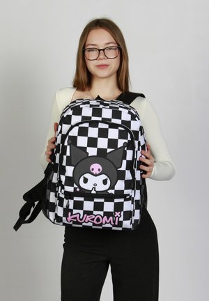 Mochila a cuadros en blanco y negro con un personaje de dibujos animados con orejas puntiagudas, un detalle rosa en la parte inferior y correas de hombro ajustables.