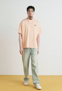 T-shirt oversized em pêssego claro com um pequeno logotipo preto, combinada com jeans verde claro e ténis brancos sobre um chão amarelo.