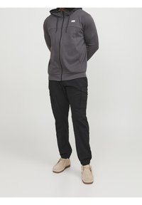 Grijze rits-hoodie met capuchon, voorzien van een borstzak; gecombineerd met zwarte cargobroeken en licht tan schoenen. Zachte textuur en casual pasvorm.
