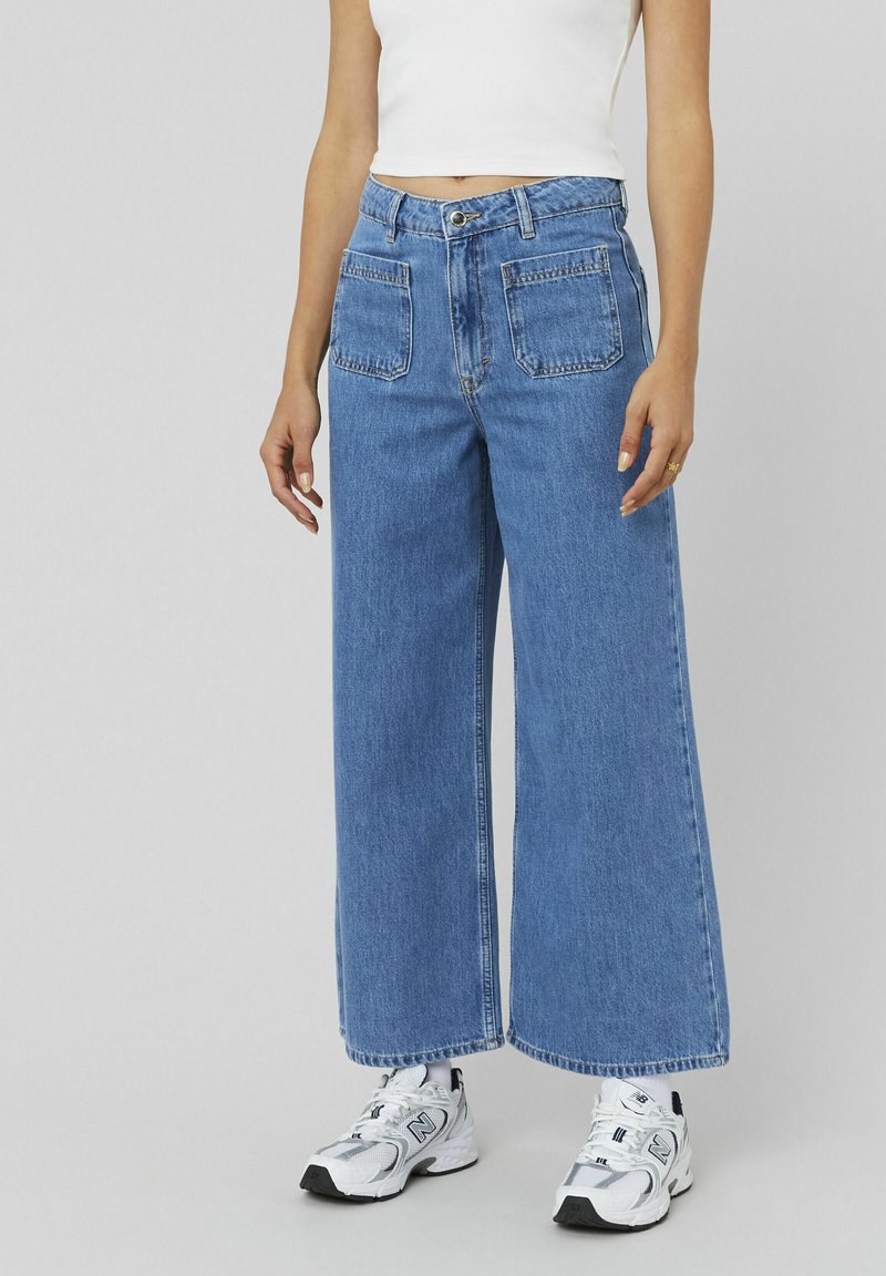 Jean large bleu clair en denim, à taille haute et avec deux poches arrière. Associé à des chaussures de sport blanches. Design simple et décontracté.