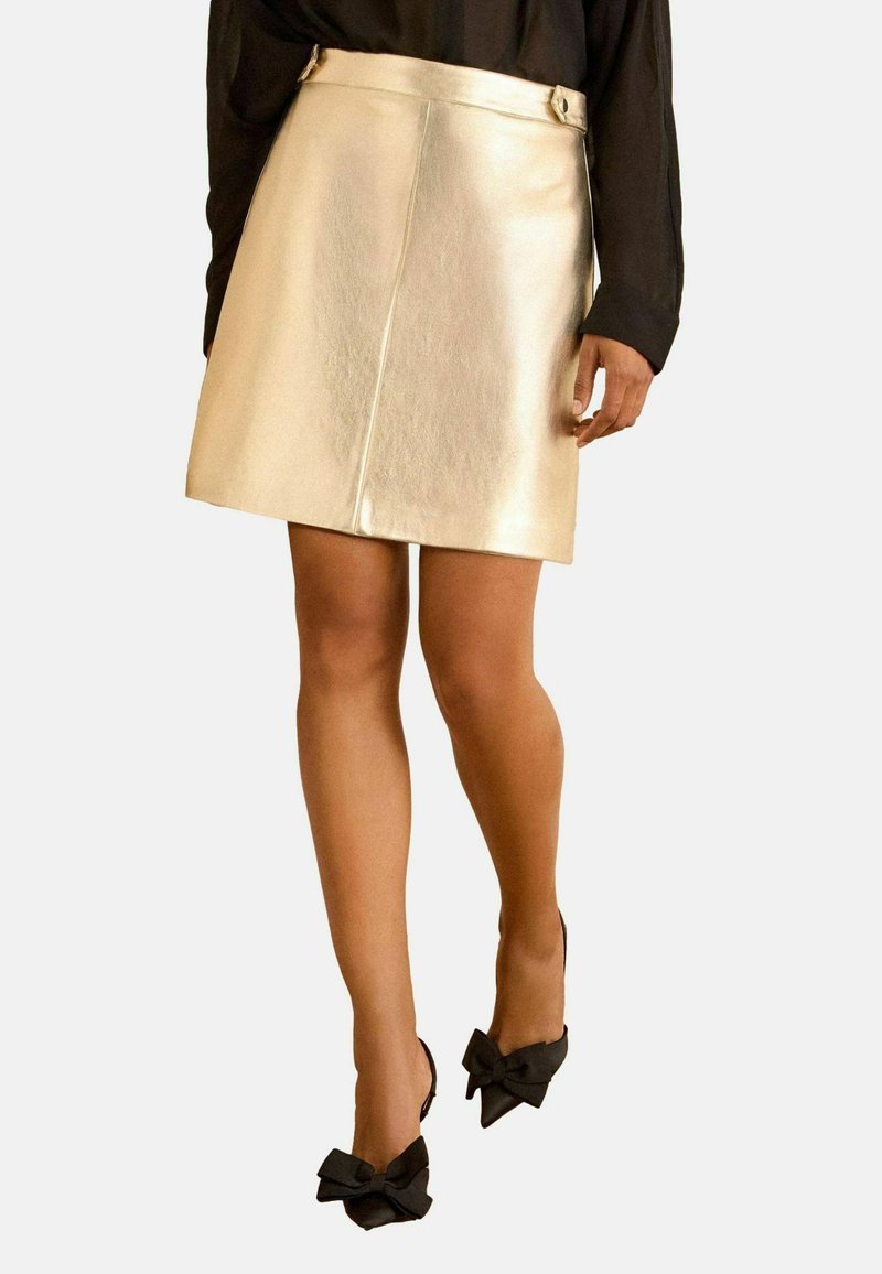 Aukso metalinė mini sijonas su lygiu paviršiumi, turintis prigludusį liemenį, šoninius mygtukus ir paprastą A formos siluetą. Juodi aukšti kulniukai su kaspinų akcentais.