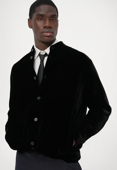 Black velvet jacket featuring a button-down front, a single chest pocket, and a relaxed fit with side pockets. Styled over a white shirt and tie.