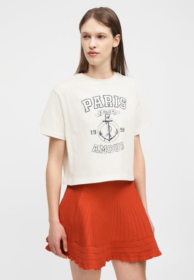Γυναίκα που φοράει λευκό κοντό μπλουζάκι με το σχέδιο «Paris Amour» και έναν άγκυρα, συνδυασμένο με κόκκινη πλισέ φούστα, σε απλό φόντο.