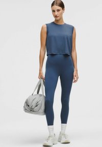 Mörkblå croppad linne och högmidjade leggings tillverkade av stretchigt material; modellen håller en ljusgrå gymväska med dubbla handtag.