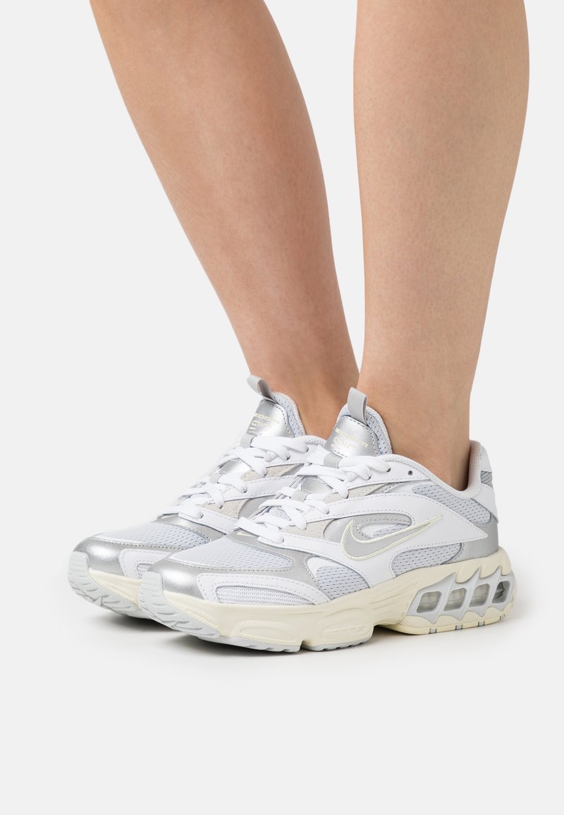 Baskets Nike blanches et argentées avec détails en mesh et cuir, portées aux pieds sur un fond clair uni.