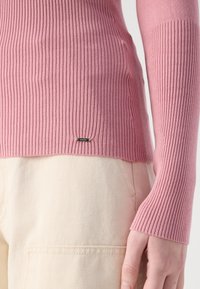 Jasnoróżowy sweter z prążkowanej dzianiny z długimi rękawami, z dyskretną metką z logo na końcu. Noszony na jasnych, fakturowanych spodniach.