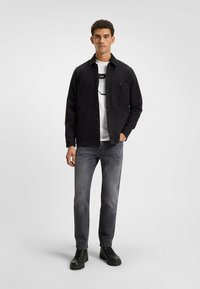 Sort skjorte med knaplukning og to brystlommer, hvid T-shirt nedenunder med et grafik, grå jeans og sorte snørebåndsstøvler.