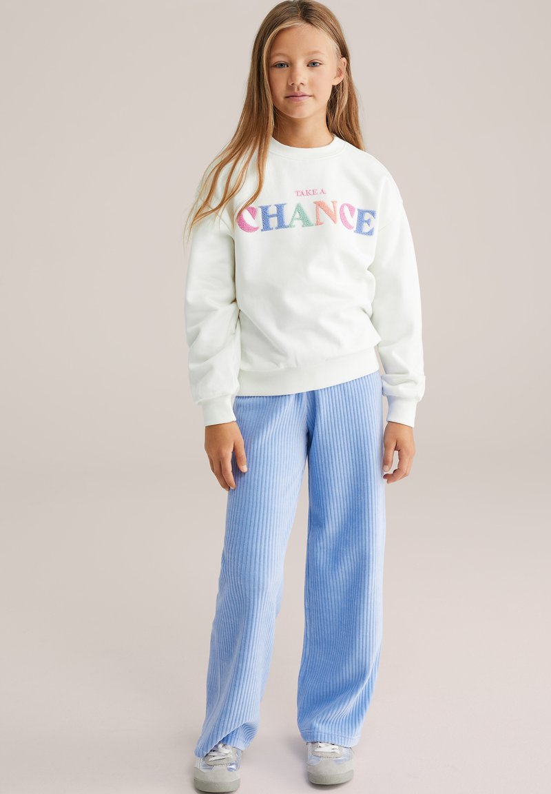 Gaiši zaļa kokvilnas krekla ar daudzkrāsainu "TAKE A CHANCE" tekstu, apvienota ar ziliem ribotiem platām biksēm, lai radītu ikdienišķu izskatu.