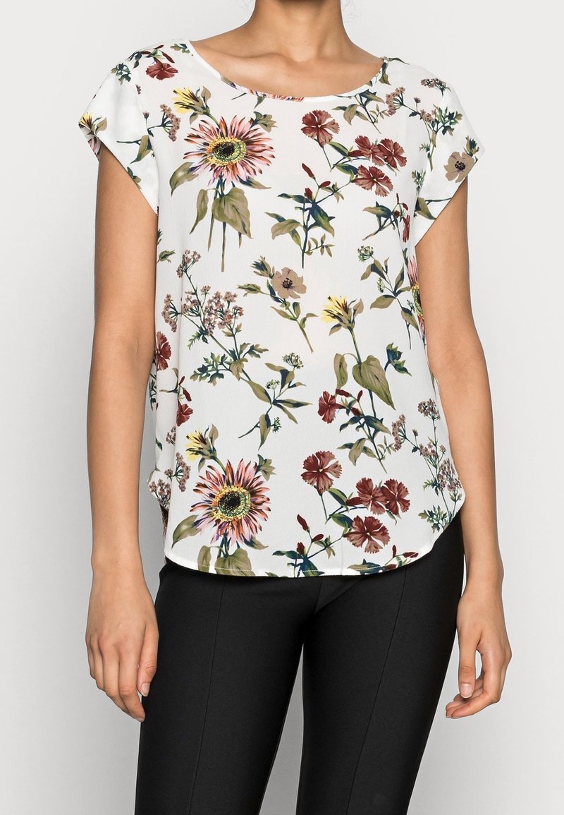 Femme portant un chemisier blanc à manches courtes avec un imprimé floral coloré et un pantalon noir ajusté, visible du cou jusqu’aux hanches.