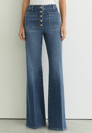 Hoch taillierte blaue Jeans mit weitem Bein, aufgesetzten Fronttaschen und fünf sichtbaren Messingknöpfen am Hosenschlitz, getragen von einer stehenden Person.