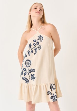 Beiges Baumwollkleid mit marineblauer floraler Stickerei, Halterneck-Design und einem Rüschensaum. Weicher Stoff, knielange Passform und lockere Schnittführung.