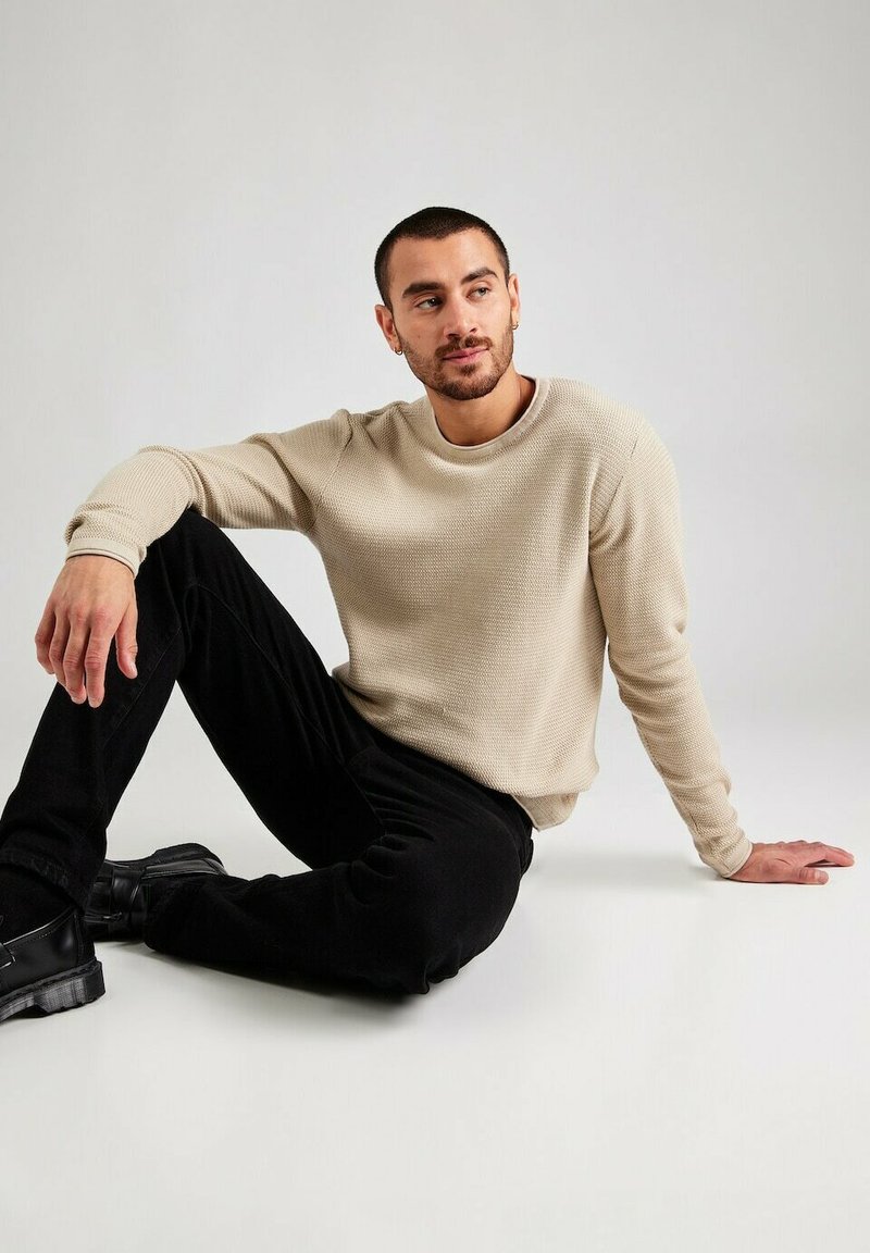 Maglione beige texturizzato con scollatura tonda, abbinato a pantaloni neri e scarpe nere robuste. Modello seduto su uno sfondo neutro.