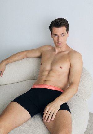 Schwarze Boxershorts mit einem roten Calvin Klein Bund, anschmiegsames Design, sitzend auf einem strukturierten hellen Stuhl vor einem neutralen Hintergrund.