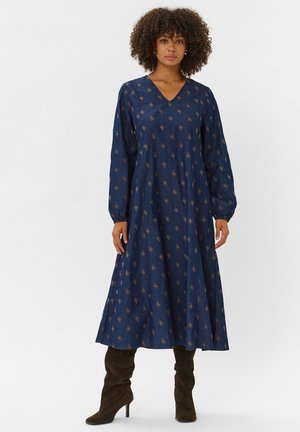 Femme aux cheveux bouclés portant une longue robe bleu marine à petits motifs floraux et des bottes hautes en daim marron sur un fond blanc uni.