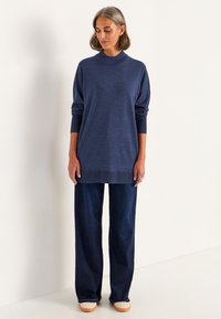 Kvinna som bär en långärmad, marinblå tröja, mörka vida jeans och vita skor, stående mot en enkel ljus bakgrund.