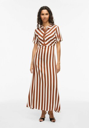 Kvinde stående iført en brun og hvid maxi kjole med lodrette striber, korte ærmer og sorte åbne hæle mod en hvid baggrund.