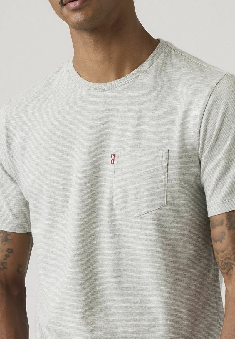Homme portant un T-shirt à manches courtes gris clair avec une poche poitrine et une petite étiquette rouge, montrant des tatouages sur les avant-bras.