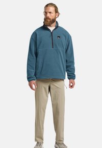 Fleece quarter-zip pullover in teal met een hoge kraag, voorzien van een zwart logo patch. Draagt een lichtbeige broek en grijze sneakers.