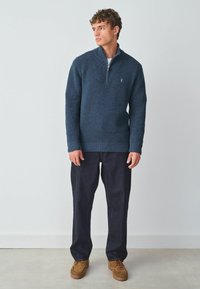 Granatowa sweter z ćwierć zamkiem, wykonany z teksturowanej tkaniny, w zestawieniu z ciemnymi dżinsami i brązowymi zamszowymi butami. Dyskretne logo na klatce piersiowej.
