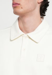 Polo bianca con colletto, finitura effetto texture, patta con due bottoni e logo ricamato sul petto.