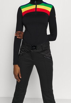 Person trägt schwarze Hose mit Reißverschluss-Details und ein schwarzes Langarmshirt mit gelben, grünen, roten und blauen Streifen auf der Brust.