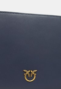 Borsa a mano in pelle blu navy con superficie texturizzata, dettagli in oro e due accenti a forma di uccello, e un anello metallico circolare.