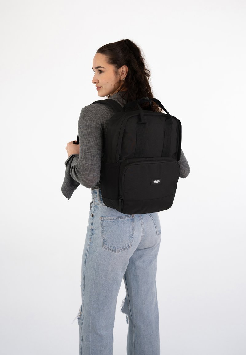 Sac à dos noir avec plusieurs compartiments, tissu robuste, forme rectangulaire, bretelles rembourrées et poche zippée à l'avant. Porté sur le dos.
