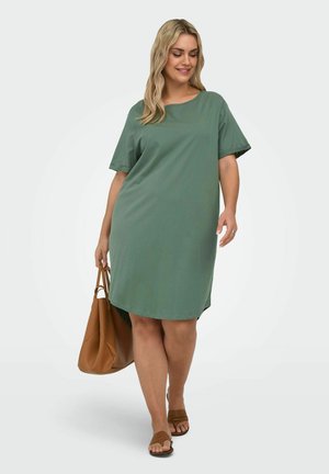 Woman wearing loose green knee-length dress, brown sandals, and carrying a large brown handbag, walking with a smile on a white background.