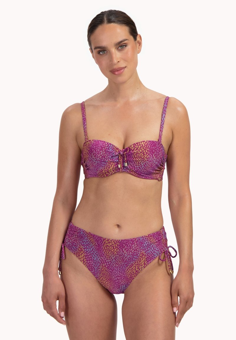 Donna che indossa un bikini viola e arancione con motivo a fantasia, con laccetti regolabili sulla parte superiore e sui lati della parte inferiore, in piedi su sfondo bianco.