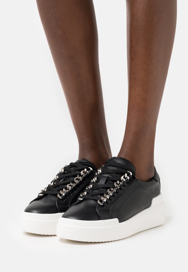 Baskets en cuir noir avec des accents de chaîne argentée et des lacets noirs, portées sur des jambes à la peau foncée sur un fond blanc uni.
