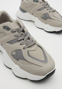 Zapatillas deportivas beige y grises con paneles de malla y ante, suelas gruesas blancas y cordones grises, situadas sobre una superficie blanca.