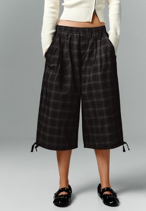 Personne portant un pantalon culotte noir à larges jambes à carreaux avec une taille élastique et des chaussures en cuir verni noir, debout les mains dans les poches.