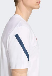 Camiseta blanca de manga corta con una franja diagonal azul marino en el hombro izquierdo y un corte relajado, fabricada con material de algodón suave.