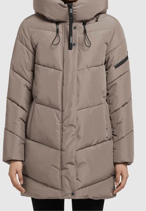 Manteau d'hiver matelassé taupe avec col montant, fermeture éclair à l'avant, boutons-pression et tirettes de fermeture éclair noires porté par une personne avec les mains relâchées sur les côtés.