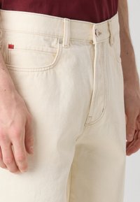 Beige puuvillahousut suoran leikkauksen omaavina. Ominaisuuksia ovat nappi- ja vetoketjusulku, vyölenkit ja pieni punainen brändilippu taskussa.