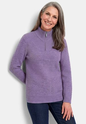 Femme aux cheveux gris portant un pull en maille violet avec fermeture éclair et un jean foncé, souriant avec une main derrière le dos.